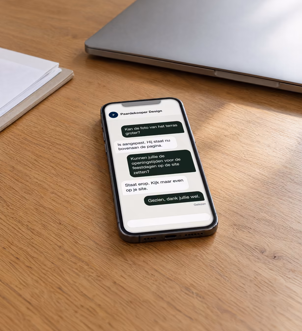 Telefoon naast een laptop op een opgeruimd bureau, overdag. Op het scherm een berichtenwisseling waarin een fictief hoveniersbedrijf een kleine wijziging aan zijn website vraagt en het antwoord krijgt dat die is aangepast