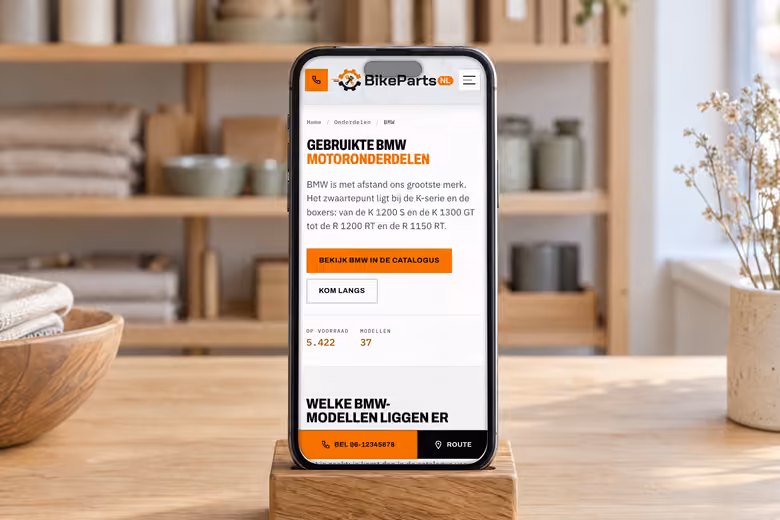 Telefoon in een eiken standaard op een lichte toonbank, overdag. Op het scherm de pagina over gebruikte BMW-motoronderdelen van BikePartsNL: een zwarte kop met een oranje regel, een oranje knop naar de catalogus en een lijst met BMW-modellen