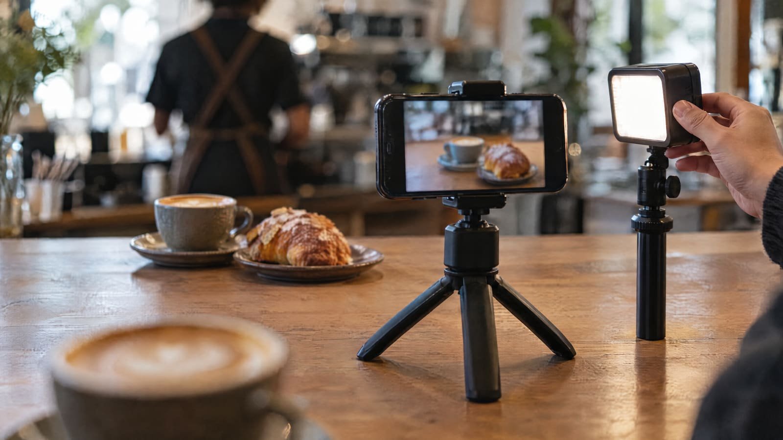 Een cafétoonbank waar een smartphone op een klein statief staat, met het scherm naar de kijker gericht en een kop koffie met gebak in beeld op het scherm; een kop koffie staat wazig op de voorgrond, terwijl een hand een klein LED-paneel bijstelt en een wazige barista op de achtergrond staat.